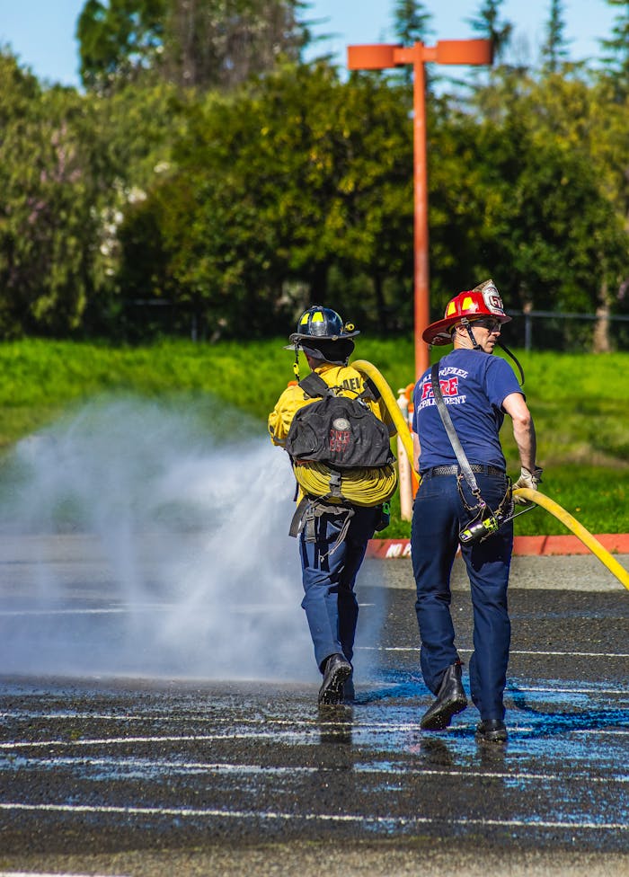 Two firefighters manage a water hose in an outdoor training session, wearing protective gear.